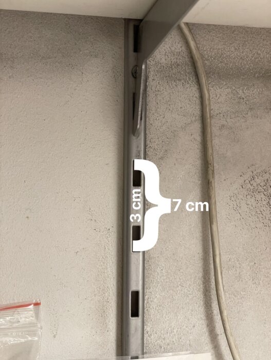 Väggskena med monterad konsoll, mätning visar 3 cm mellan fästhål och 7 cm mellan skruvhål, kabel synlig till höger.