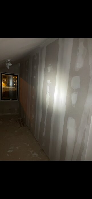 Nymålad innervägg med synliga vertikala linjer och ojämn spackling mellan gipsskivor, belyst med ficklampa i ett mörkt rum.