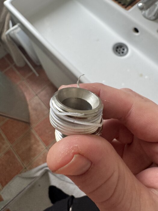 En person håller en excenterkoppling med packningstråd för blandarfäste. I bakgrunden syns ett handfat.