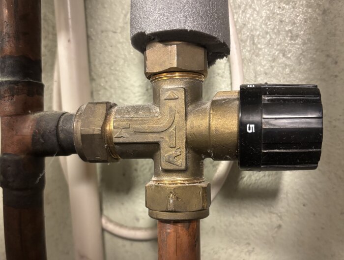 En mässingsblandningsventil med svart vred med siffror synlig, ansluten till rör i en fjärrvärmecentral, visad från sidan.