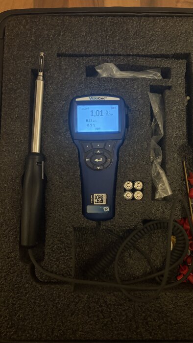 En digital anemometer med display och sond i ett vadderat fodral, använd för att mäta luftflöde och temperatur.