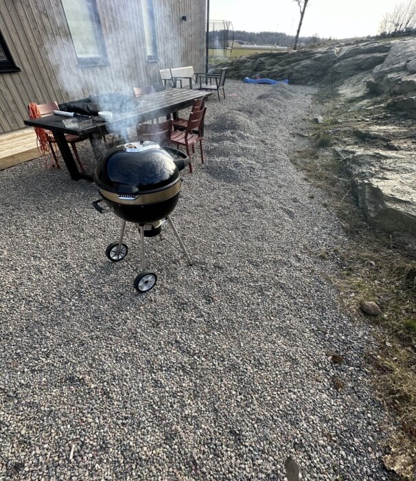 Grill med rök på grusyta vid husvägg, bord och stolar i bakgrunden, stengrund och bergkant synliga.