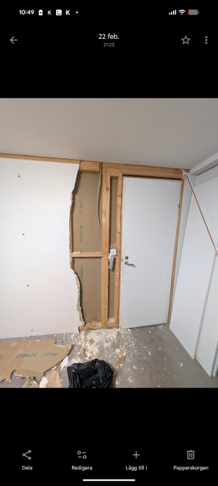 Riven gipsvägg i garage, träreglar synliga, rester på golvet, dörr till höger.