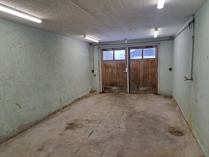 Källargarage med betonggolv och slitna träportar, ljusgröna väggar med märken, tak med lysrör, golvbrunn i hörnet.
