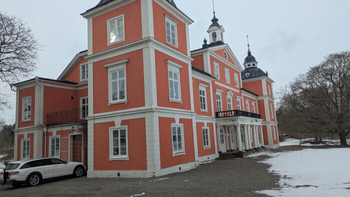 Stor röd herrgård med flera våningar, vita fönster och en grusig uppfart, omgiven av snötäckt mark och träd.