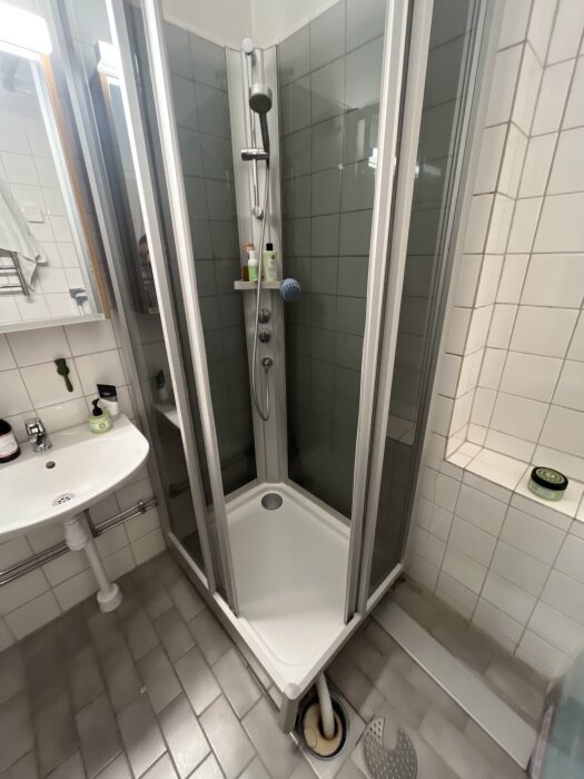 Duschhörna i ett kaklat badrum med klinkergolv, duschslang, hylla med flaskor, handfat med tvålförpackning, avloppsbrunn och hål i golvet.