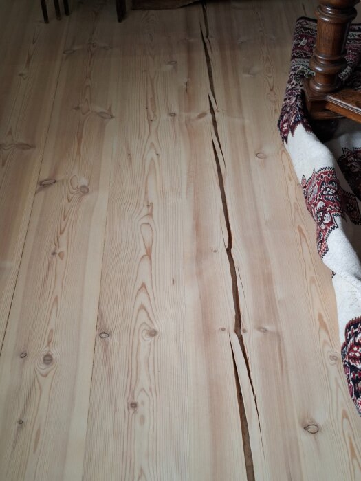 Massivt trägolv med en lång spricka mitt på, delvis täckt av en dekorativ matta i hörnet.