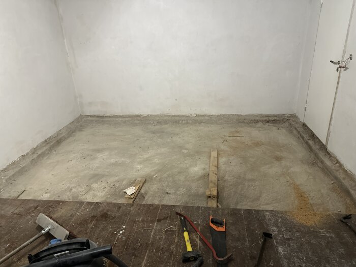 Rum med betonggolv som förbereds för renovering. Verktyg som såg och hammare samt träbitar syns på golvet.