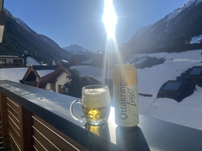 En ölflaska och ett glas öl på en balkong med snötäckta tak och berg i bakgrunden under en solig dag.