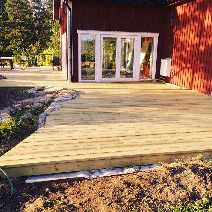 Nybyggd träaltan vid rött hus med stora glasdörrar, omgiven av stenar och grönska i bakgrunden.
