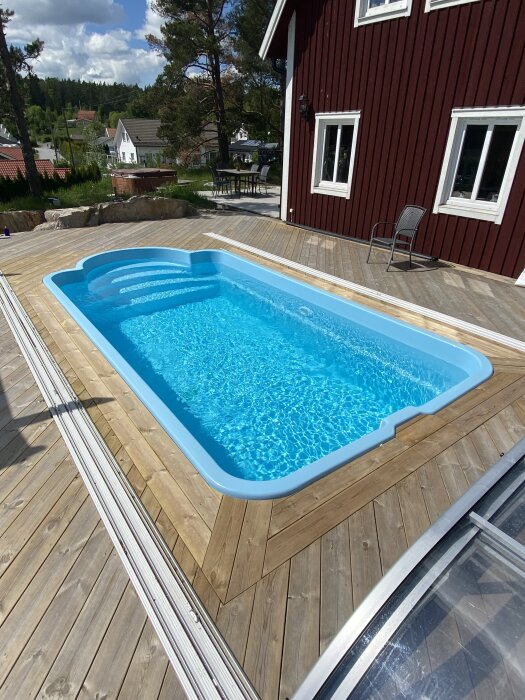 Altan med en inbyggd pool, omgiven av trädäck och nära ett rött hus. Träden i bakgrunden skapar en naturskön miljö.