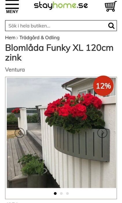 Blomlåda i zink fylld med röda blommor hänger på en vit vägg vid en altan, med sjö i bakgrunden. Rabatt på 12% visas på bilden.
