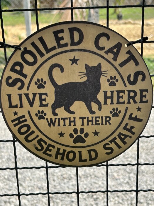 Skylt med texten "Spoiled Cats Live Here With Their Household Staff" och en bild av en katt, hänger på ett nätstaket.