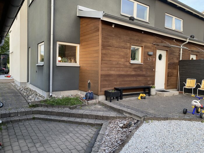 Framsidan av ett hus med trappsteg och stenlagd yta, kantad av stenar och grus. Två stolar och en barnleksak syns också.