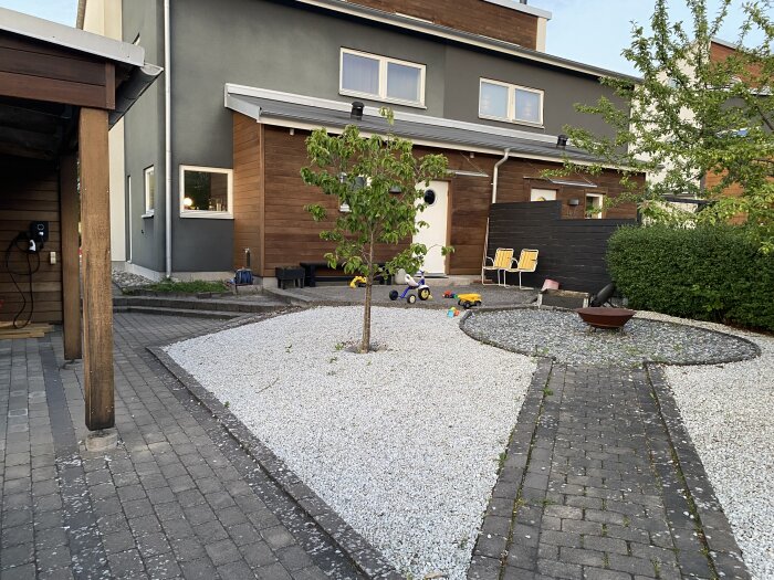 Framsida av hus med grus och stenbeläggning, en synlig trädstam, leksaker, sittplatser, samt en blandning av asfalt och träpanel på husväggen.