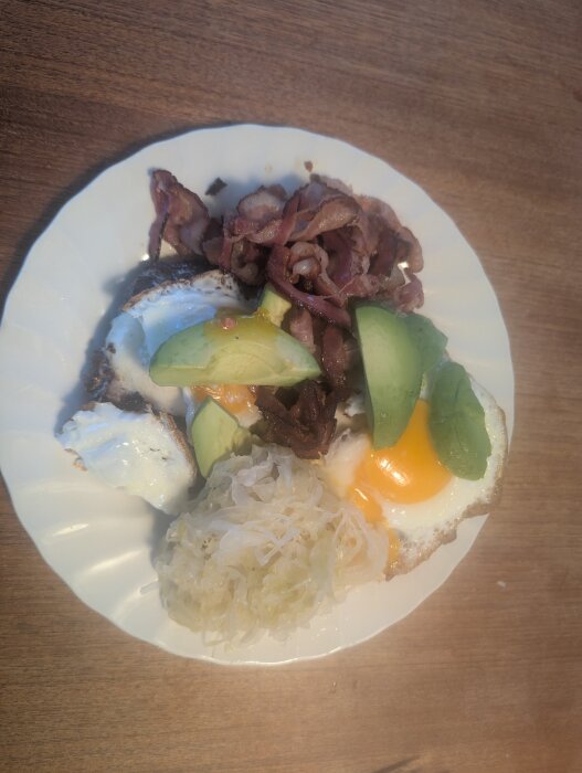 Tallrik med bacon, stekt ägg med rinnig gula, avokado och surkål.