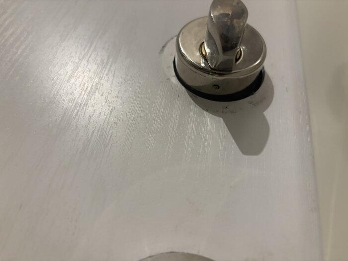 Lockdel av blank metall på vit dörr, delvis borttagen med synlig skruv. Används för att byta låscylinder.