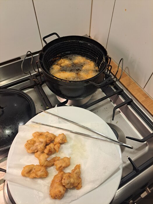 Friterad kyckling i en korg över en gryta med olja på spisen; flera bitar ligger på en tallrik med papper och tång.
