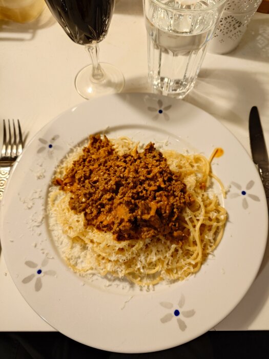 Spagetti med köttfärssås på en tallrik, strödd med riven ost. Röda vink glas och vattenglas är placerade bredvid, med bestick bredvid tallriken.