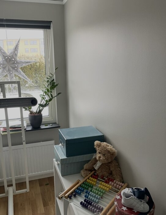 Ljust rum med nymålad vägg och fönster. Hylla med nallebjörn, kulram och förvaringslådor. Barnsko och pyssel papper på fönsterbräda.
