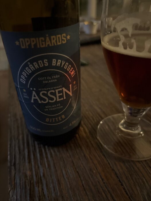 Flaska med etikett från Oppigårds Bryggeri samt glas med mörk öl på ett träbord.