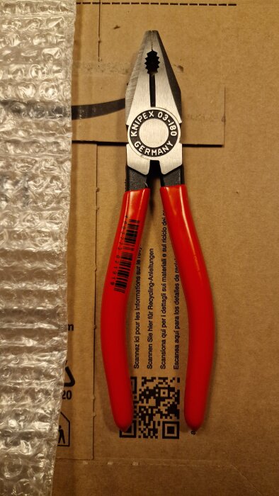 Tänger med röda handtag på en kartong, bredvid bubbelplast. Märket visar "Knipex 03-180 Germany".