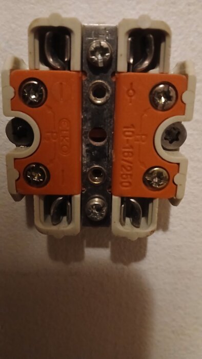 Orange keramisk kopplingsplint med metallskruvar monterad på en vägg. Texter och symboler synliga på ytan.