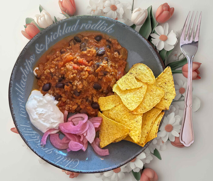 Chili con carne med svarta bönor och chorizofärs, serverad med picklad rödlök, gräddfil och tortillachips på en tallrik med blommönster.