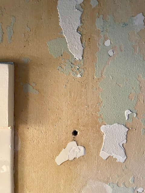 Närbild av köksvägg med avflagnande tapet som blottar betongliknande yta, samt ett hål i väggen.