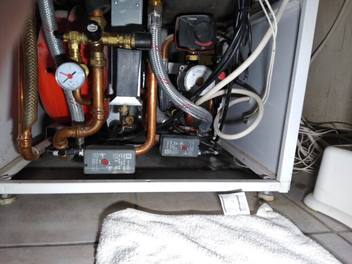 Översiktsbild av inre komponenter i en varmvattenberedare med rör, kopplingar och en mätare; läckage nära en pump syns på golvet.