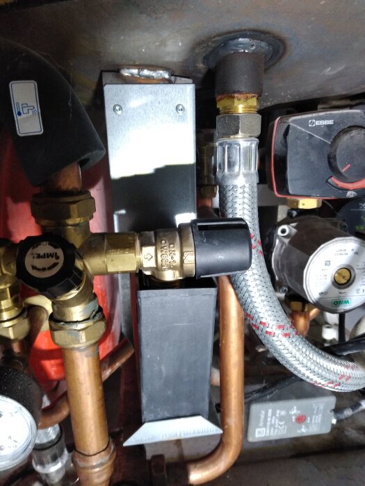 Rörinstallation med säkerhetsventil, flera kopparrör och en flexibleslang kopplad till en ventil i en metallstruktur, möjligen del av ett värmesystem.