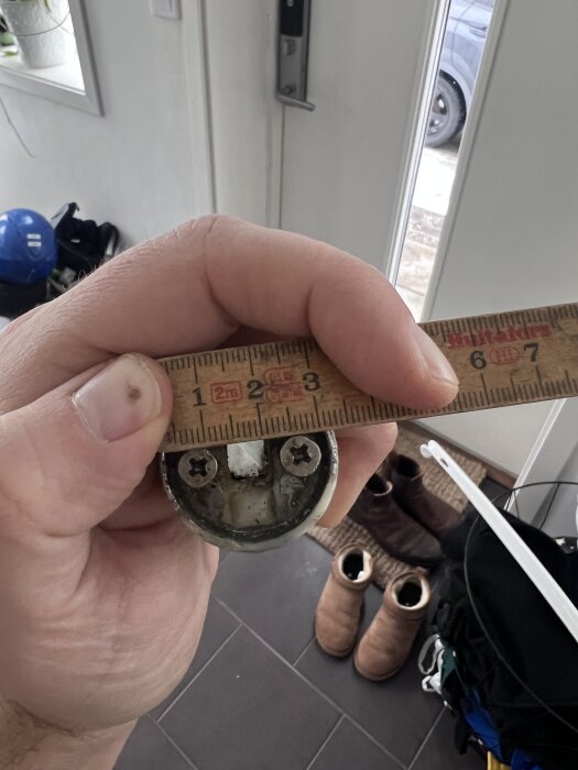 En hand håller en rund metallfäste med två skruvar och ett måttband placerat över det, inomhus med skor och kläder i bakgrunden.