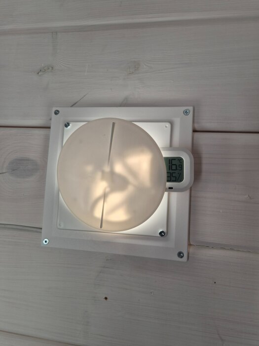 Solventilationssystem monterat på vägg i friggebod, rund täckplåt med skåra, digital termometer visar 16,9 grader.