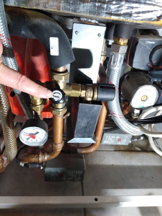 En person pekar på en temperaturegulator bland rör och ventiler i ett värmesystem. Det finns även en tryckmätare och rörkopplingar.