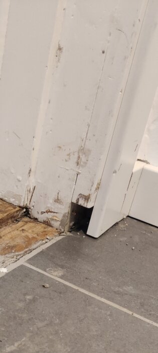 Skadat dörrkarmshörn nära golvet, delvis flisat trä och smutsigt, bredvid grått kakelgolv med vit foglinje.