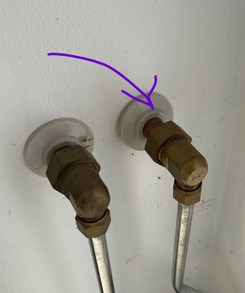 Två kopparrör med mässingsfittings anslutna till en vägg, markerade med lila pil, pekar på ett eventuellt skadat område vid ena rörets anslutning.