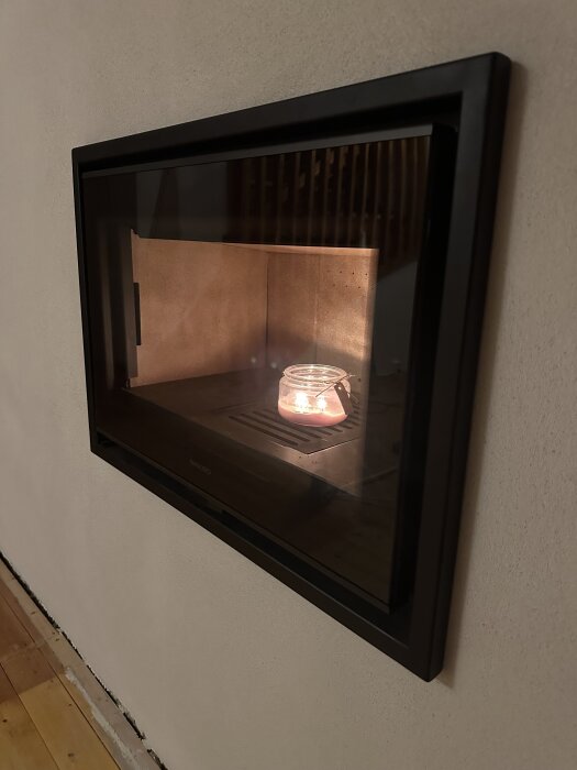 Inbyggd kamin i en vägg med ett tänt ljus inuti, nyligen putsad yta och trägolv i gillestuga under renovering.