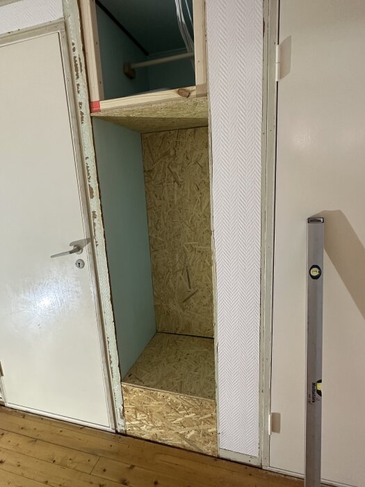 Öppen väggkonstruktion med OSB-skivor i nyrenoverad källare, intill vit dörr och nivåmätare på trägolv.