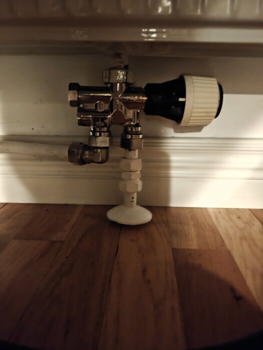 Termostatventil och rörkopplingar under ett element, sett mot en träparkettbakgrund.