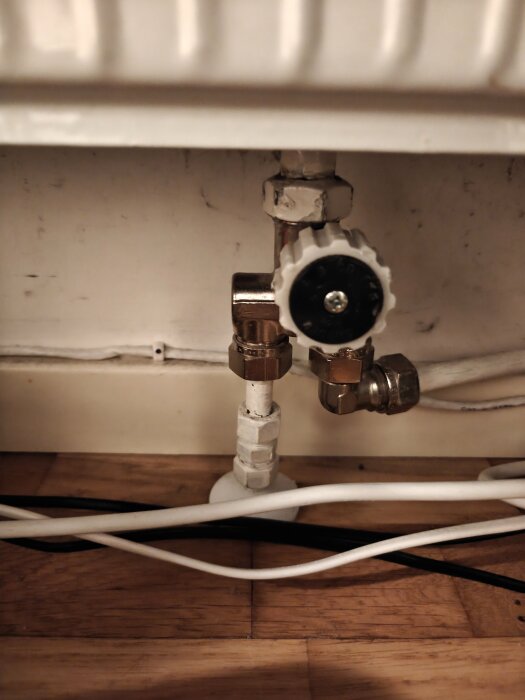 Närbild av en vit radiatorventil under ett element, med synliga kopparrör och elkablar på trägolv i bakgrunden.