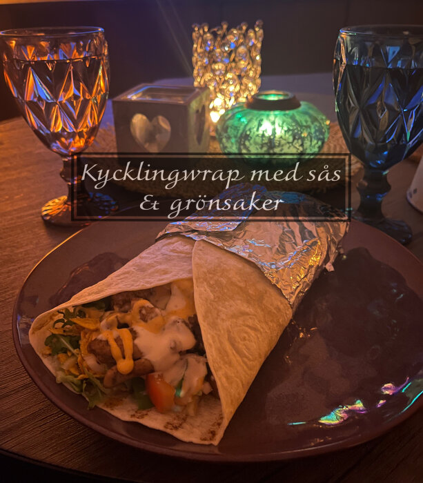 Kycklingwrap med sallad, ruccola, picklad rödlök, kebabsås, tomat och gurka, serverad på en tallrik med dricksglas och ljuslyktor i bakgrunden.