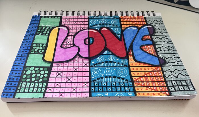 Texten "LOVE" i färgglad blockstil, omgiven av mosaikmönster i olika färger och former på ett spiralblock.