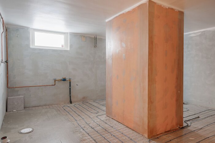 Ofärdigt rum med stengolv, väggar av putsad betong och synliga vattenrör. Golvet har värmeslingor; ett stort, färgat betongblock står centralt.