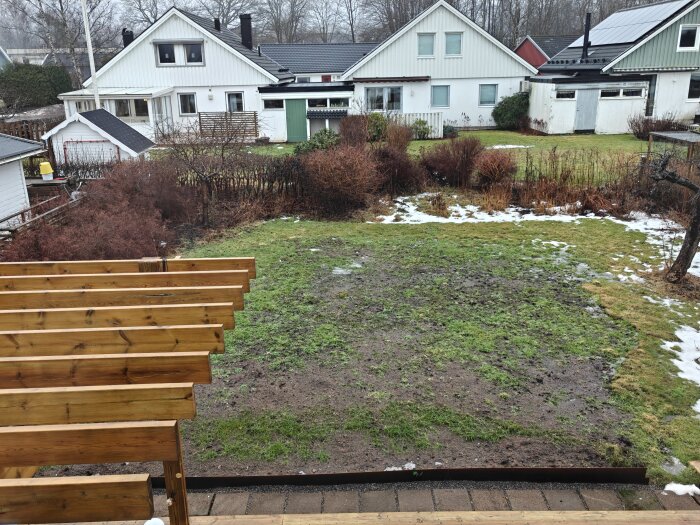 Gles, grön gräsmatta med bar jord och fläckar av snö, omgiven av buskar och flera villor i bakgrunden. I förgrunden syns en träpergola.