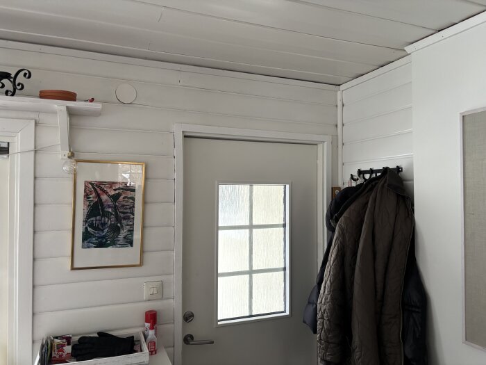 Entré med vitmålad träpanel, ytterdörr med fönster, kapphängare med jackor och en tavla på väggen.
