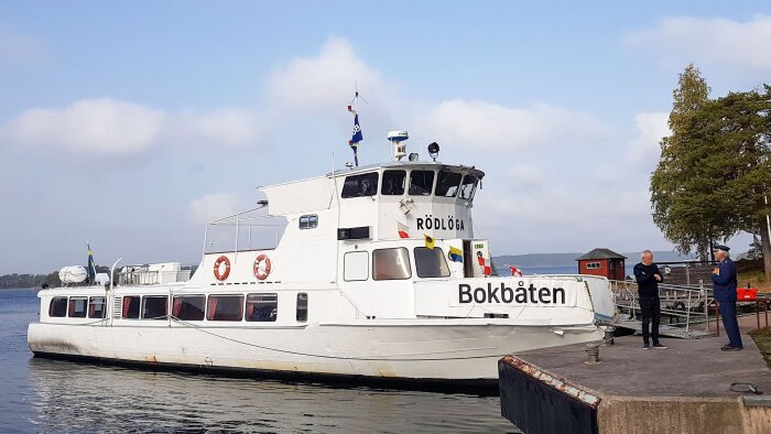 En vit passagerarbåt benämnd "Bokbåten" vid en brygga, med två personer i samtal bredvid.