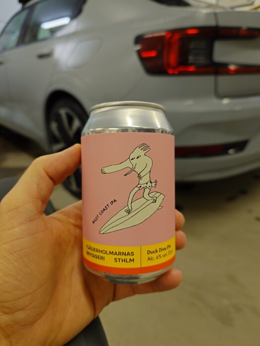 En hand håller en burk West Coast IPA från Fjäderholmarnas Bryggeri, med en bil i bakgrunden.