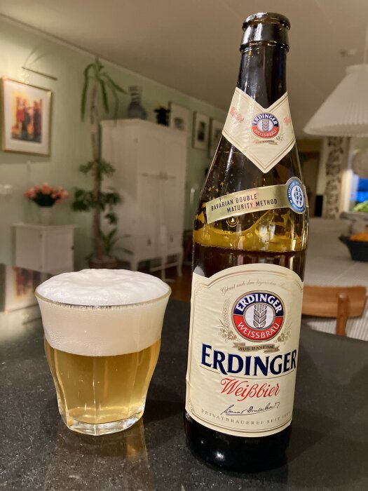 Ölglas med skum bredvid en flaska Erdinger Weißbier på ett köksbord.