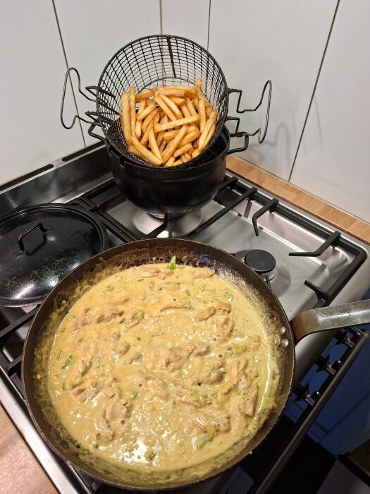 Stekpanna med krämig kycklinggryta på spis och fritös med pommes frites i korg ovanpå.