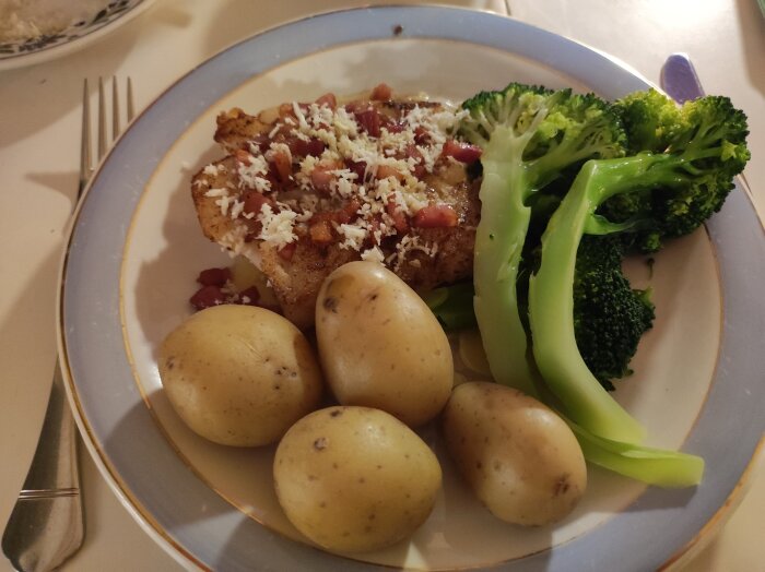 Tallrik med stekt fisk toppad med bacon och riven ost, omgiven av tre kokta potatisar och broccoli.